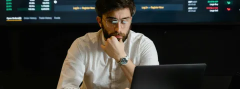 Un señor están mirando el ordenador y por detrás hay gráficos de finanzas en una pantalla