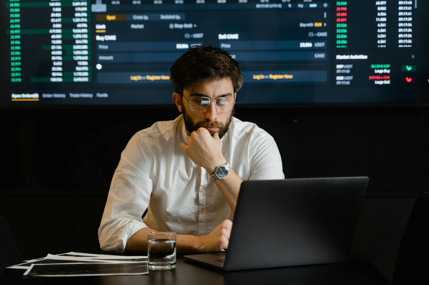 Un señor están mirando el ordenador y por detrás hay gráficos de finanzas en una pantalla