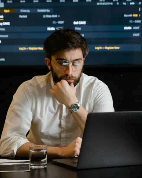 Un señor están mirando el ordenador y por detrás hay gráficos de finanzas en una pantalla